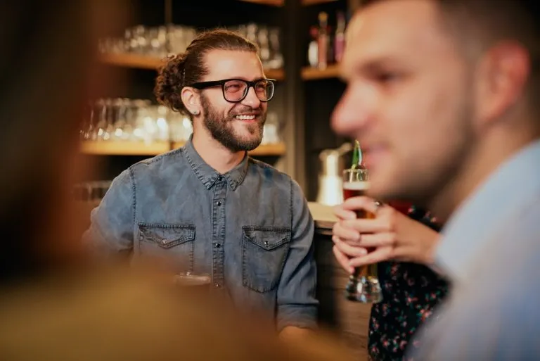 Gut aussehender, lächelnder Hipster-Mann, der in einem Pub in der Nähe der Theke sitzt, ein Bier in der Hand hält und interessante Geschichten hört. Nachtleben.