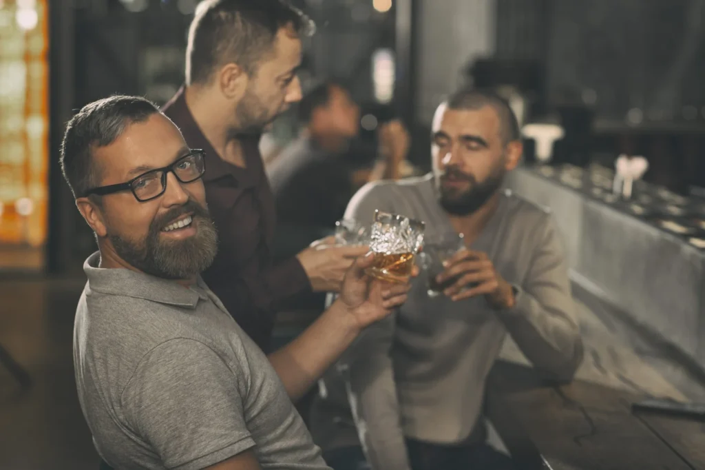 Fröhlicher bärtiger Mann hält ein Kristallglas mit Whisky in der Hand, schaut in die Kamera und lächelt. Die Kunden der Bar sitzen an der Theke dahinter, unterhalten sich und trinken Alkohol.